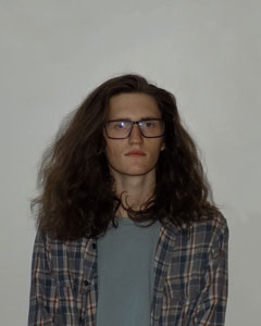 a portrait of Jackson Huckaby. a clean-shaven white person with glasses and long hair sitting facing the camera, portrait cuts off at sternum height.